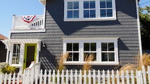 A two-story house with a white picket fence, a green door, and a balcony with a flag. - Charming modern bungalow w/ hot tub steps away from the beach (Dillon Beach)