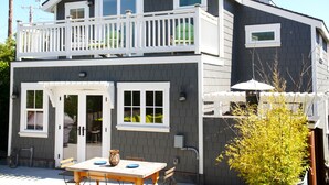 A two-story house with a balcony, a front door, and a small patio area with a table and chairs. - Charming modern bungalow w/ hot tub steps away from the beach (Dillon Beach)
