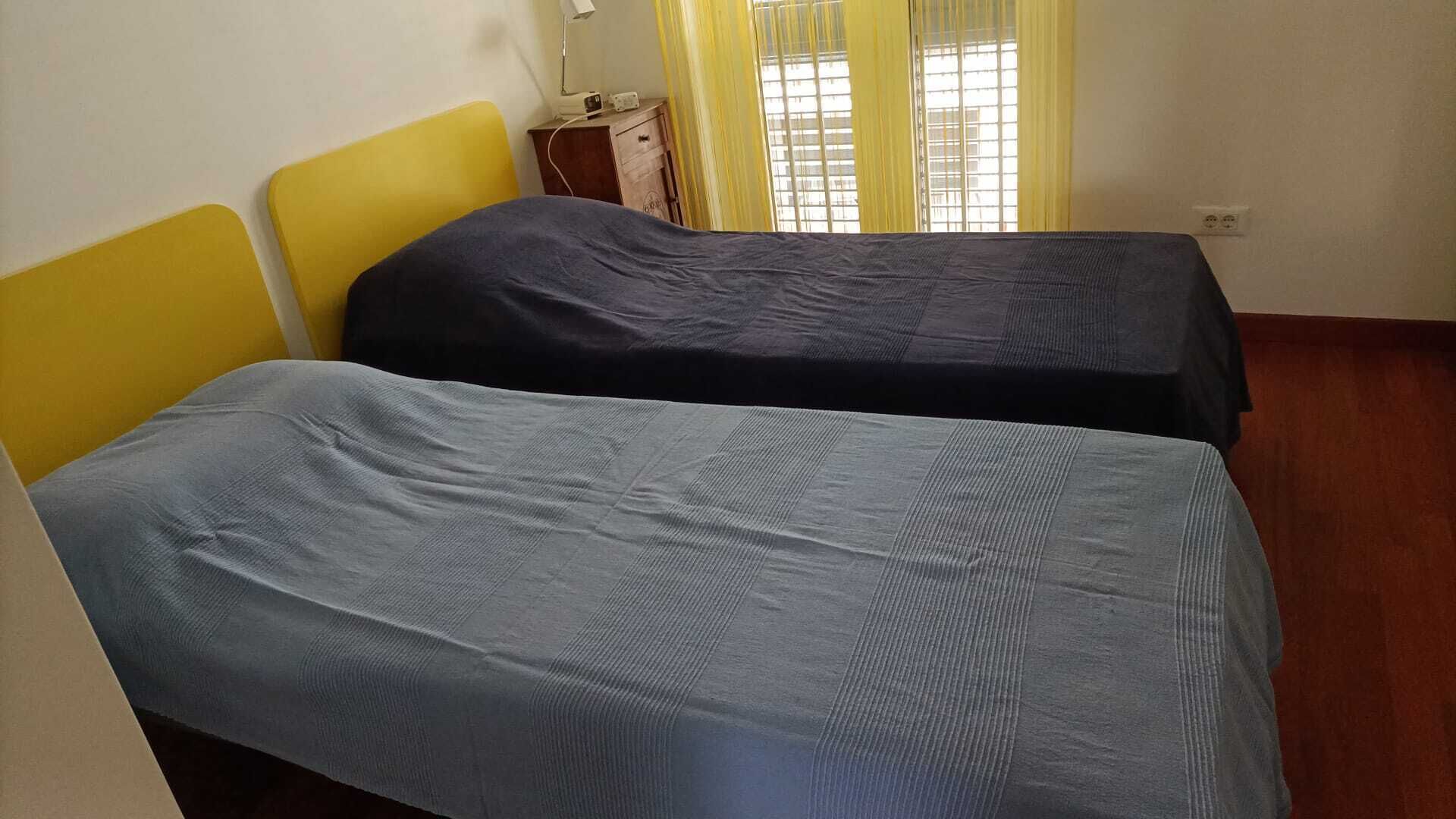 Una stanza con due letti singoli, un comodino in legno con una radio e una finestra con tende gialle.