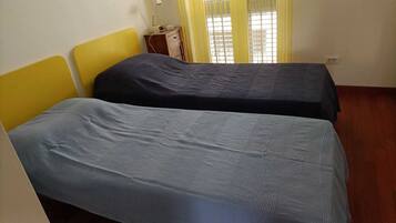Una stanza con due letti singoli, un comodino in legno con una radio e una finestra con tende gialle.