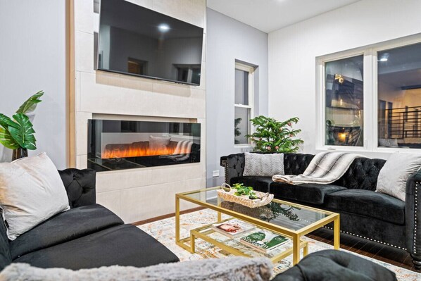 Una sala de estar moderna con chimenea, sofás negros, una mesa de centro de vidrio y un ventanal grande.