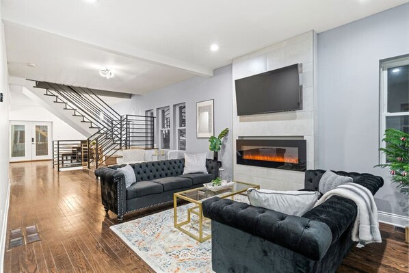 A modern living room with a fireplace, a staircase, and a large TV. - Stunning & Spacious 7BD Home with garage! (Chicago)