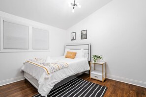 A bedroom with a bed, a bedside table, a nightstand, a window with blinds, and a ceiling light fixture.