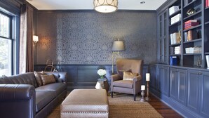 A room with a dark blue patterned wallpaper, a leather sofa, a matching ottoman, a wooden side table with a vase, a tall armchair, a floor lamp, and a bookshelf filled with books. - Edwards House (Fort Collins)