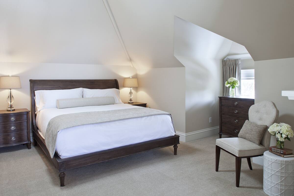 Une chambre à coucher avec un grand lit, deux tables de chevet, une commode, une chaise et une petite table d’appoint sur laquelle se trouve un vase.