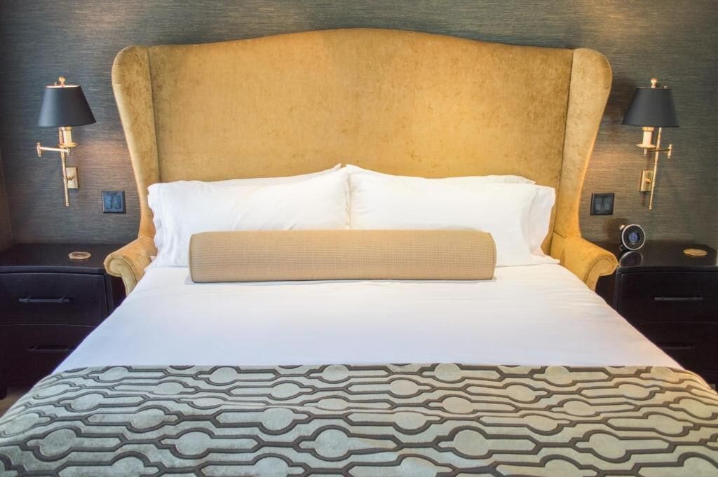 A hotel room with a large bed, two bedside tables, a headboard, and wall-mounted lamps.