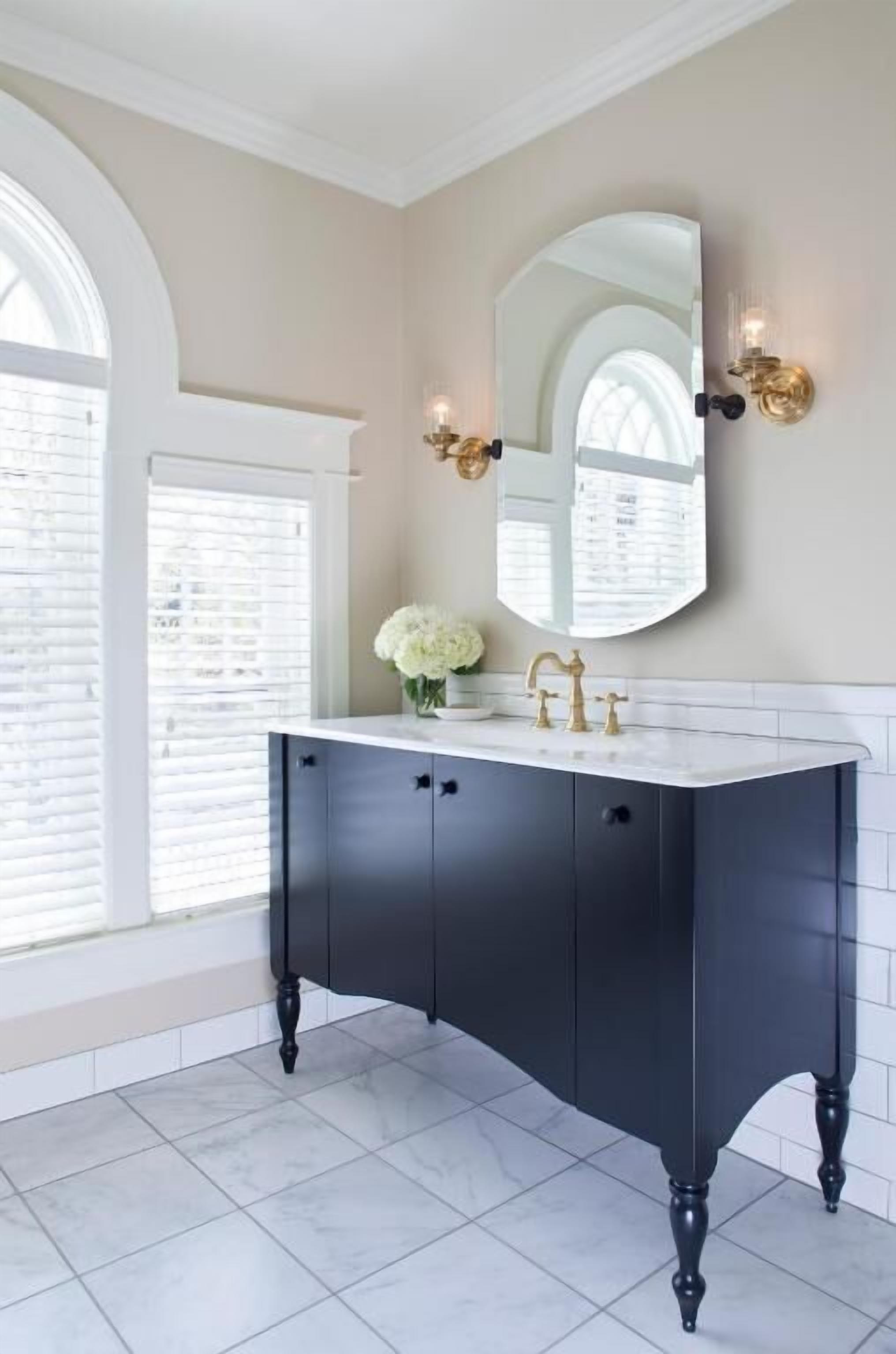 Un baño con tocador oscuro, lavamanos blanco, grifo dorado y un espejo grande con apliques de pared.