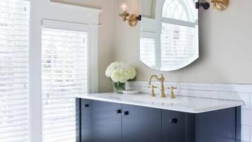 Un baño con tocador oscuro, lavamanos blanco, grifo dorado y un espejo grande con apliques de pared.
