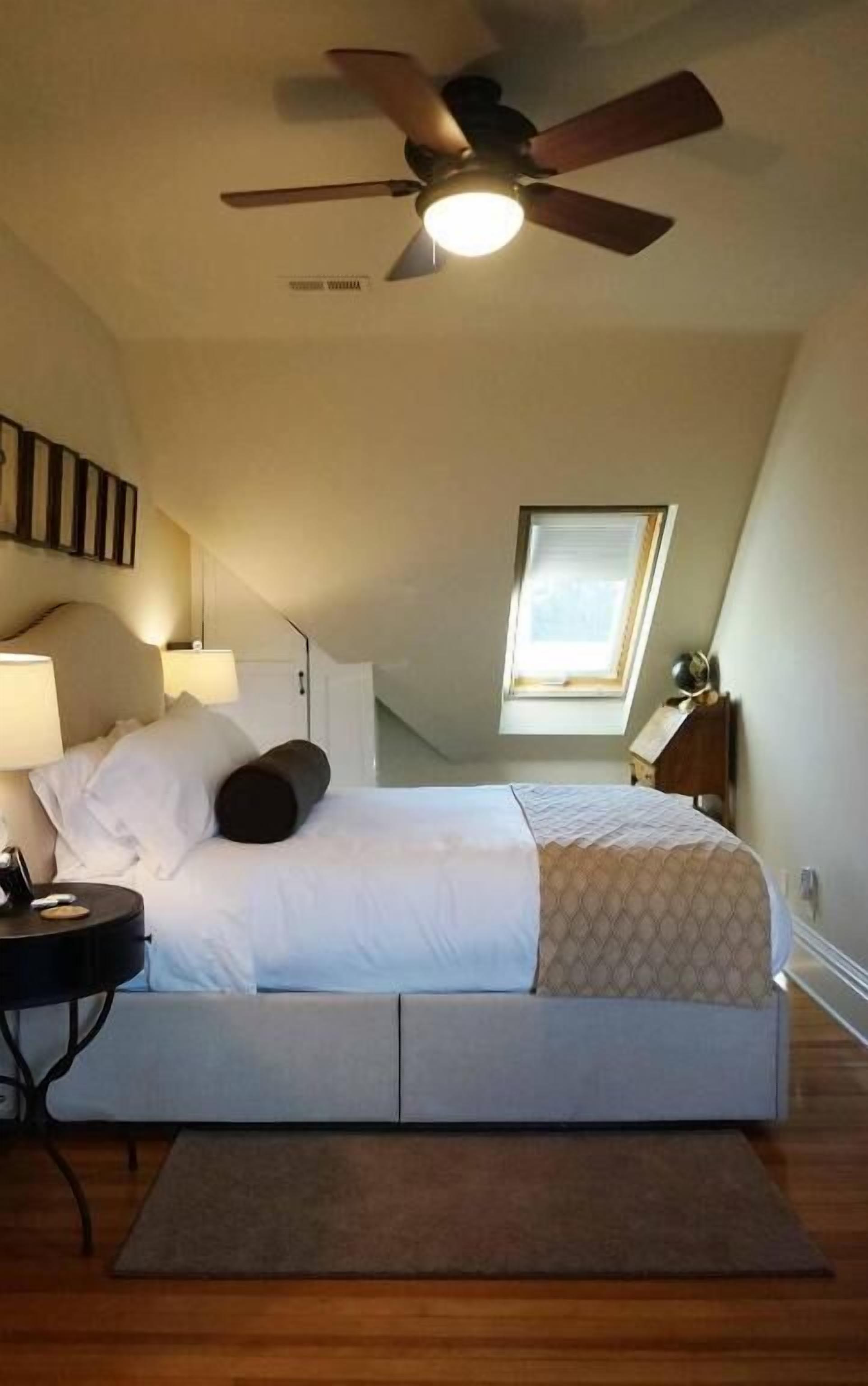 A bedroom with a bed, a nightstand, a desk, a ceiling fan, and a window.