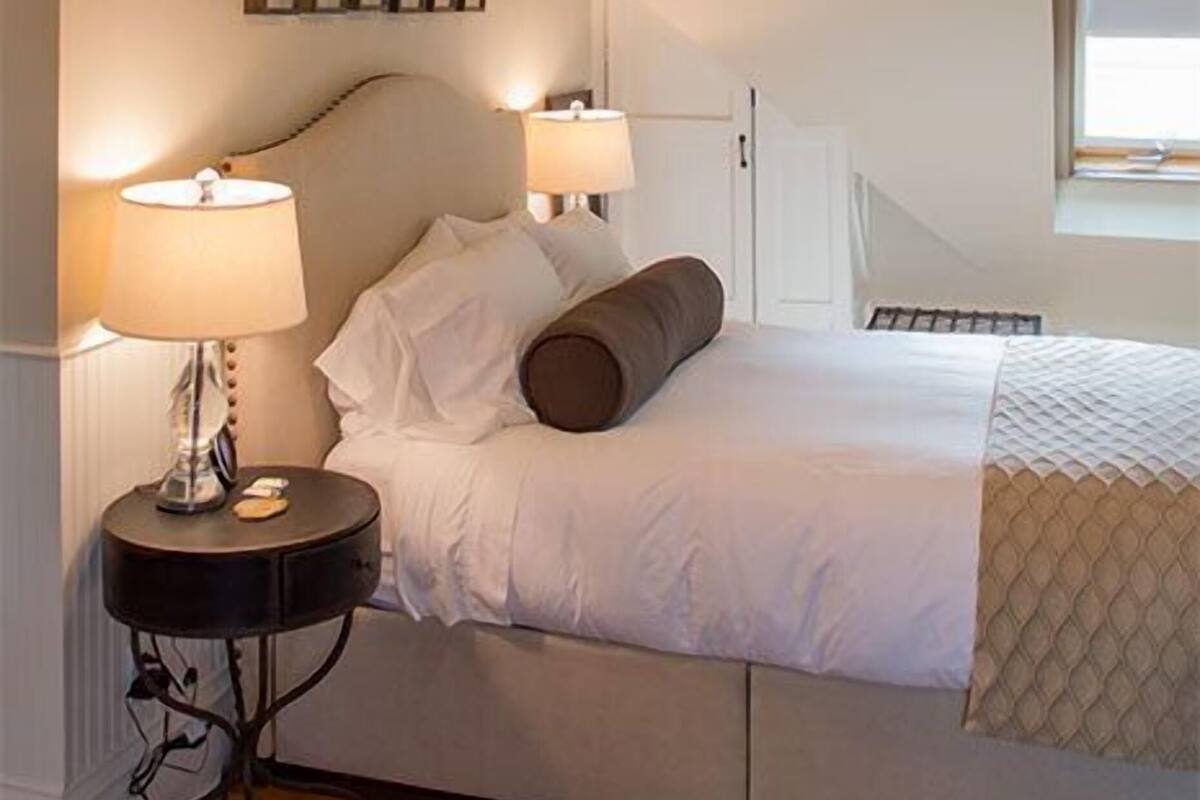 Une chambre à coucher avec un lit, deux lampes de chevet, un ventilateur de plafond, une petite table ronde et une fenêtre.