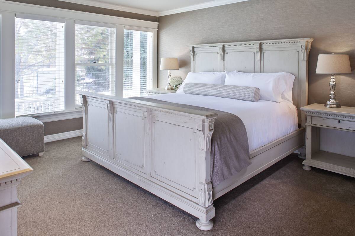 Une chambre à coucher avec un grand lit, deux tables de chevet avec des lampes, une fenêtre avec des stores et un banc recouvert d’un tissu gris.