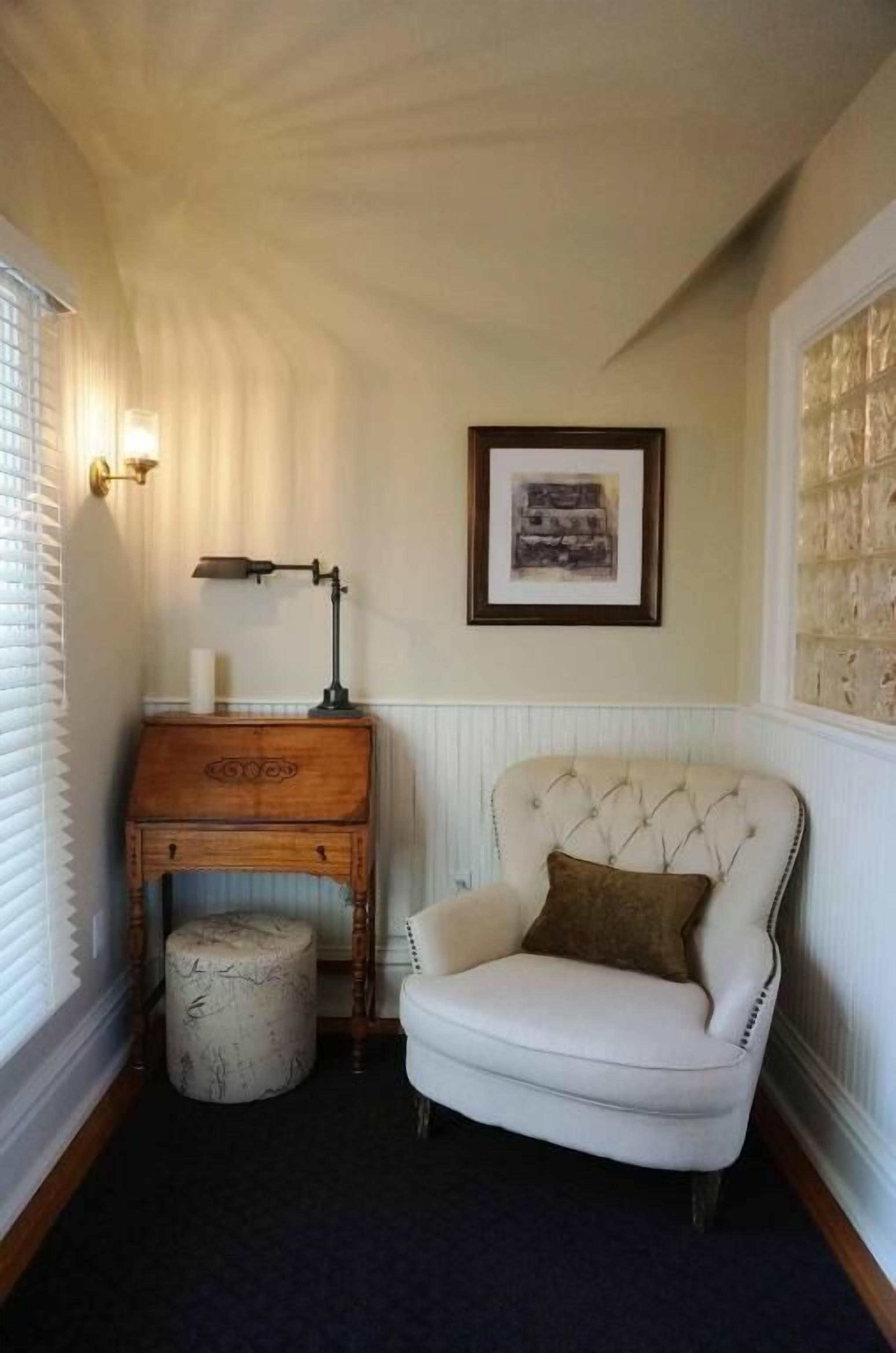 A corner room with a white armchair, a wooden chest of drawers, a lamp, and a framed picture on the wall.