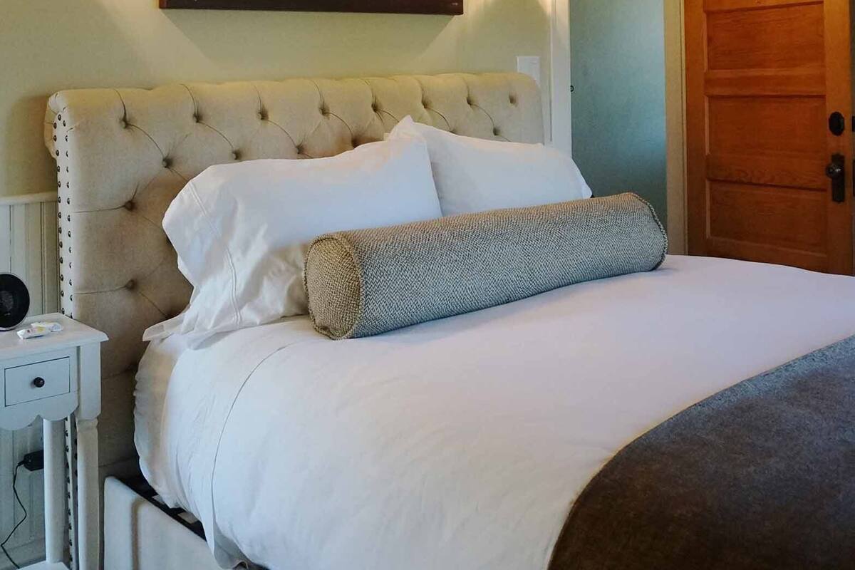 Une chambre à coucher avec une tête de lit capitonnée, un lit recouvert de linge de lit blanc, un tableau accroché au mur, une lampe de chevet et une porte en bois.