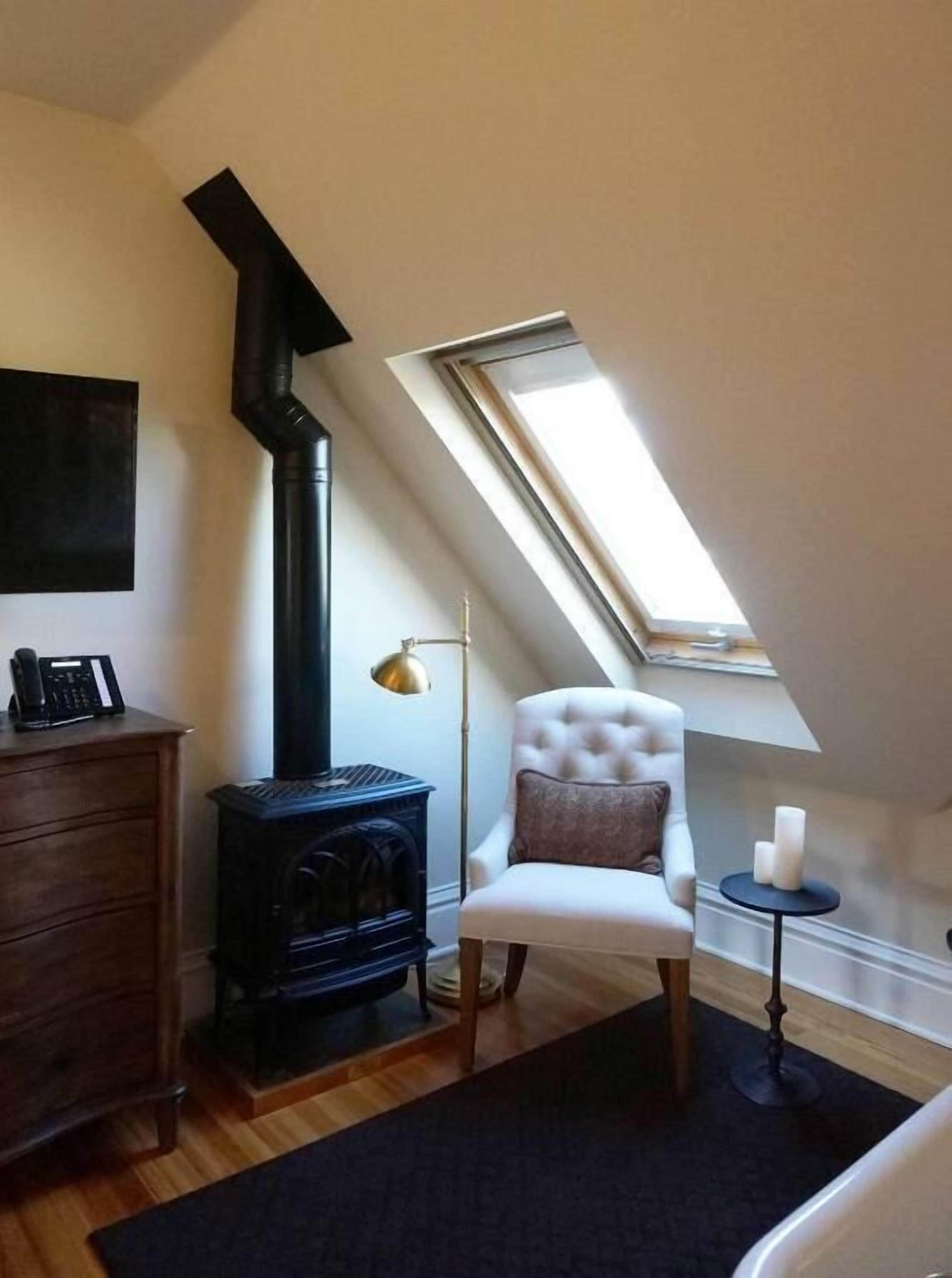 A cozy room with a black wood-burning stove, a white upholstered chair, a small round table with candles, and a slanted ceiling skylight.
