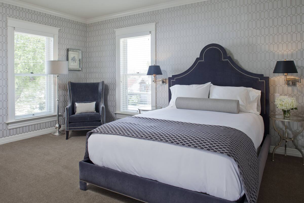 Une chambre à coucher avec un grand lit, deux tables de chevet avec des lampes, une chaise et une fenêtre avec des stores.