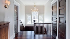 A hallway with a wooden floor, a chandelier, and a view of a window with a door on the right. - Edwards House (Fort Collins)
