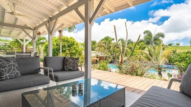 An outdoor patio with a glass-top table, two grey sofas with patterned cushions, and a view of a pool and tropical vegetation.
