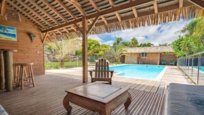 A wooden deck with a pool, a thatched-roof structure, and a wooden table.