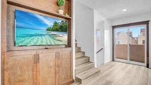 Una habitación amplia con una gran televisión de pantalla plana montada en un mueble de madera, una escalera de madera y un cerramiento de vidrio corredero que da a un área exterior.
