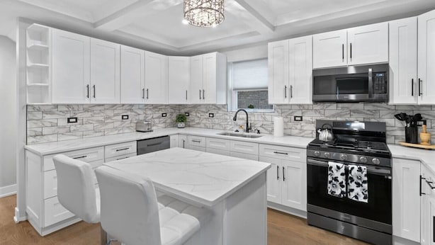A modern kitchen with white cabinetry, a marble backsplash, a central island with chairs, a stainless steel stove, and a microwave.