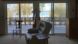 Ein Zimmer mit einer großen Schiebeglasdoor, die auf eine Terrasse mit Blick auf Bäume und ein Gewässer führt. Es gibt einen Ledersessel und einen Tisch mit einer Lampe.