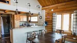A wooden cabin interior with a kitchen, dining area, and large windows.