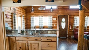 A log cabin interior with a kitchen area featuring wooden cabinets, granite countertops, and pendant lighting.