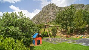Un'area giochi con altalene e una piccola struttura rossa e blu, immersa nel verde lussureggiante con una montagna sullo sfondo.