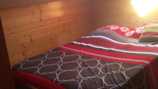Una habitación pequeña y acogedora con techo inclinado y paredes de madera. Cuenta con una cama con colcha y almohadas estampadas, y un tragaluz sobre la cama.