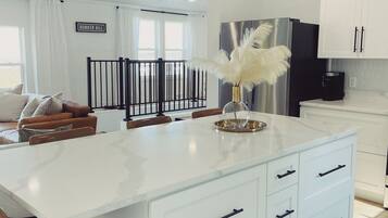 A modern kitchen with a large island, white cabinetry, black pendant lights, and a view of the outdoors through large windows.