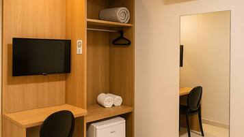 Una habitación de hotel compacta con un escritorio de madera, una silla negra, un lavavajillas pequeño blanco, un televisor montado en la pared, un espejo y una alfombra estampada.