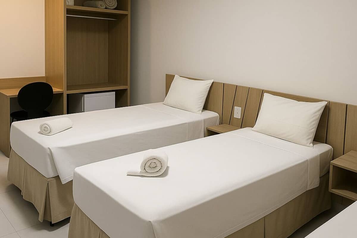 A hotel room with two single beds, each with a white bedspread and a rolled towel. There is a wooden wardrobe and a small wooden nightstand with a lamp.