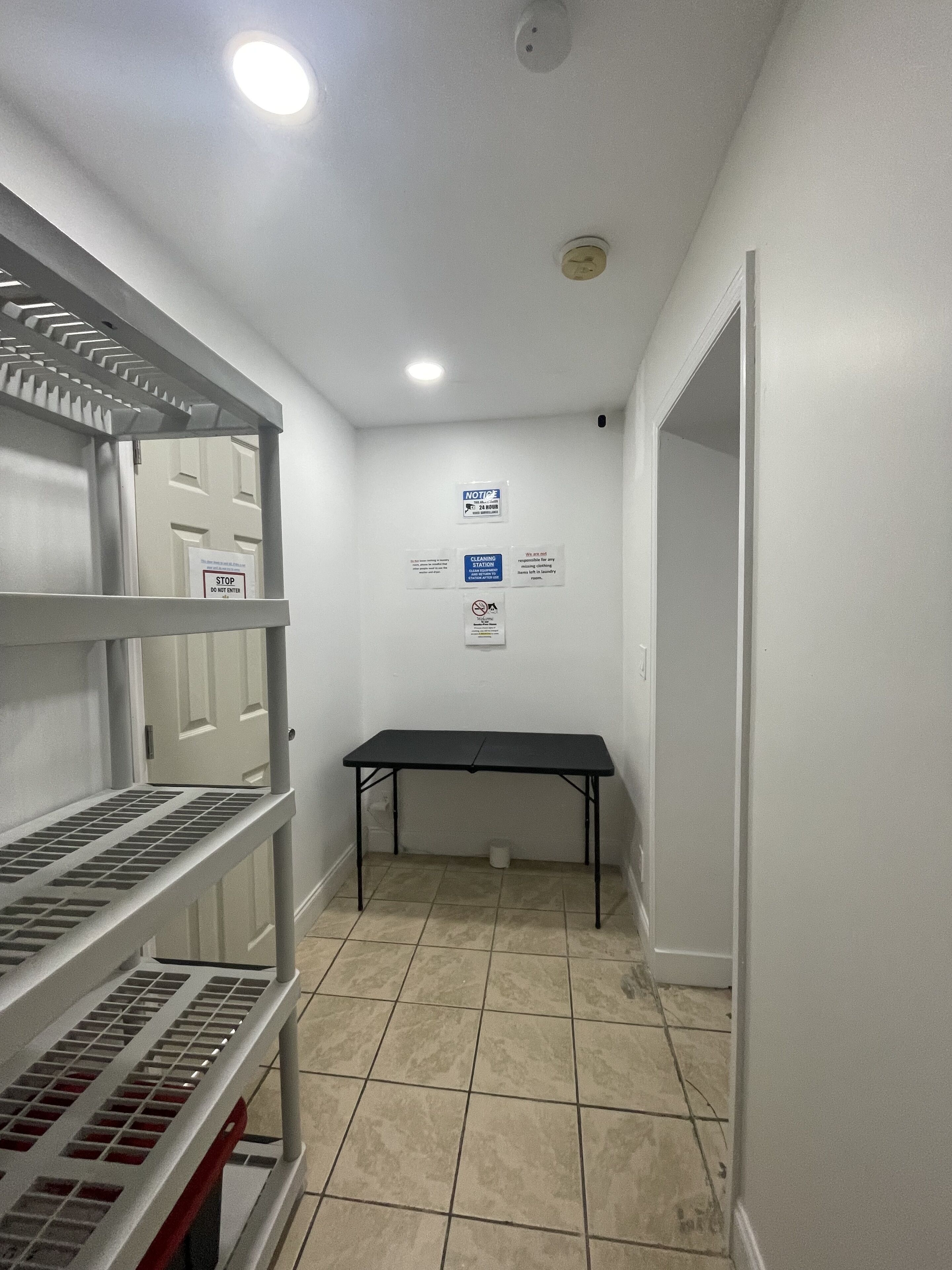 Un pasillo con una puerta blanca, una mesa negra y estantes con contenedores de almacenamiento. Hay carteles en la pared que indican instrucciones de limpieza y un aviso.