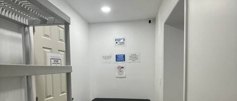 Un pasillo con una puerta blanca, una mesa negra y estantes con contenedores de almacenamiento. Hay carteles en la pared que indican instrucciones de limpieza y un aviso.
