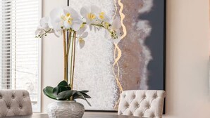 Un comedor con una mesa redonda, dos sillas tapizadas, un jarrón con flores blancas y un cuadro abstracto de gran tamaño en la pared.