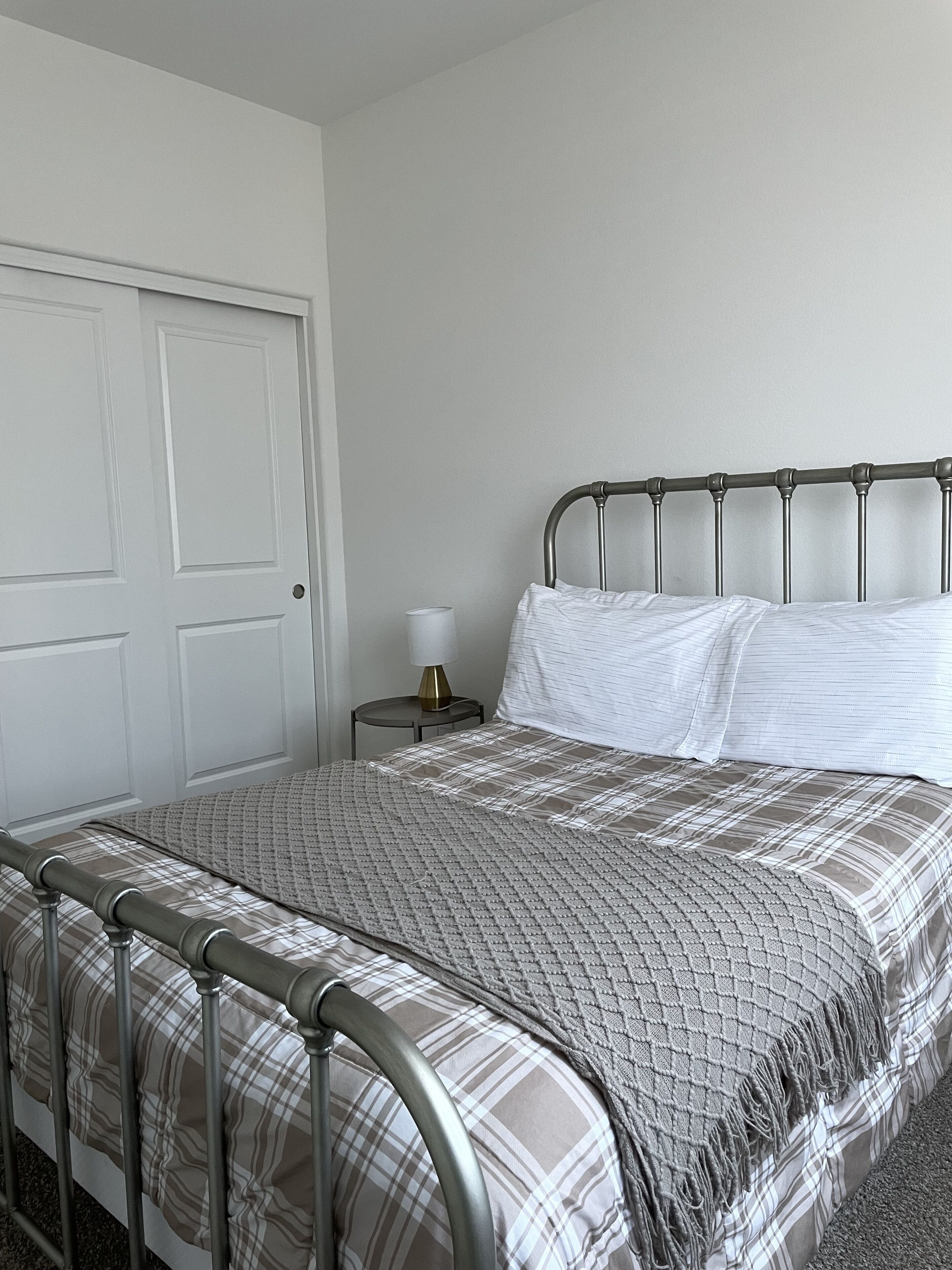 A bedroom with a metal bed frame, a plaid bedspread, a white pillow, and a bedside table with a lamp.