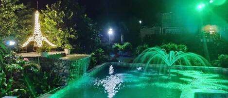 Vista nocturna de una piscina con una fuente, rodeada de vegetación y una réplica iluminada de la Torre Eiffel.