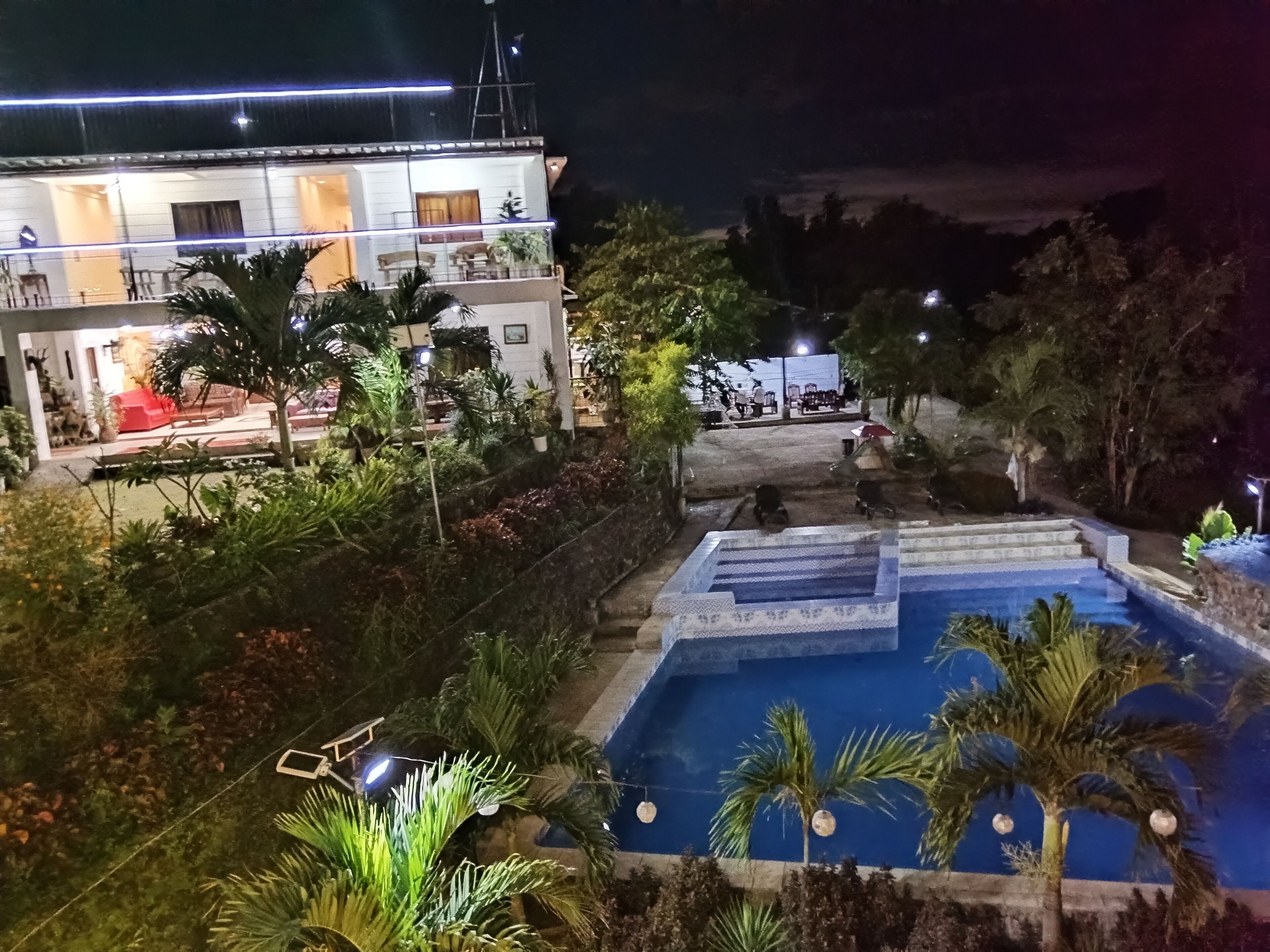 Une vue nocturne d’un complexe hôtelier avec une piscine, une végétation luxuriante et un bâtiment doté d’un espace extérieur aménagé pour s’asseoir.