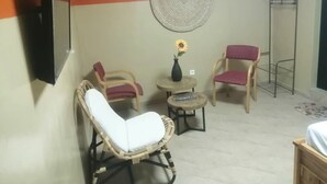 Una habitación con una televisión montada en la pared, dos sillones de mimbre, una mesita redonda con un jarrón de flores y un perchero metálico.