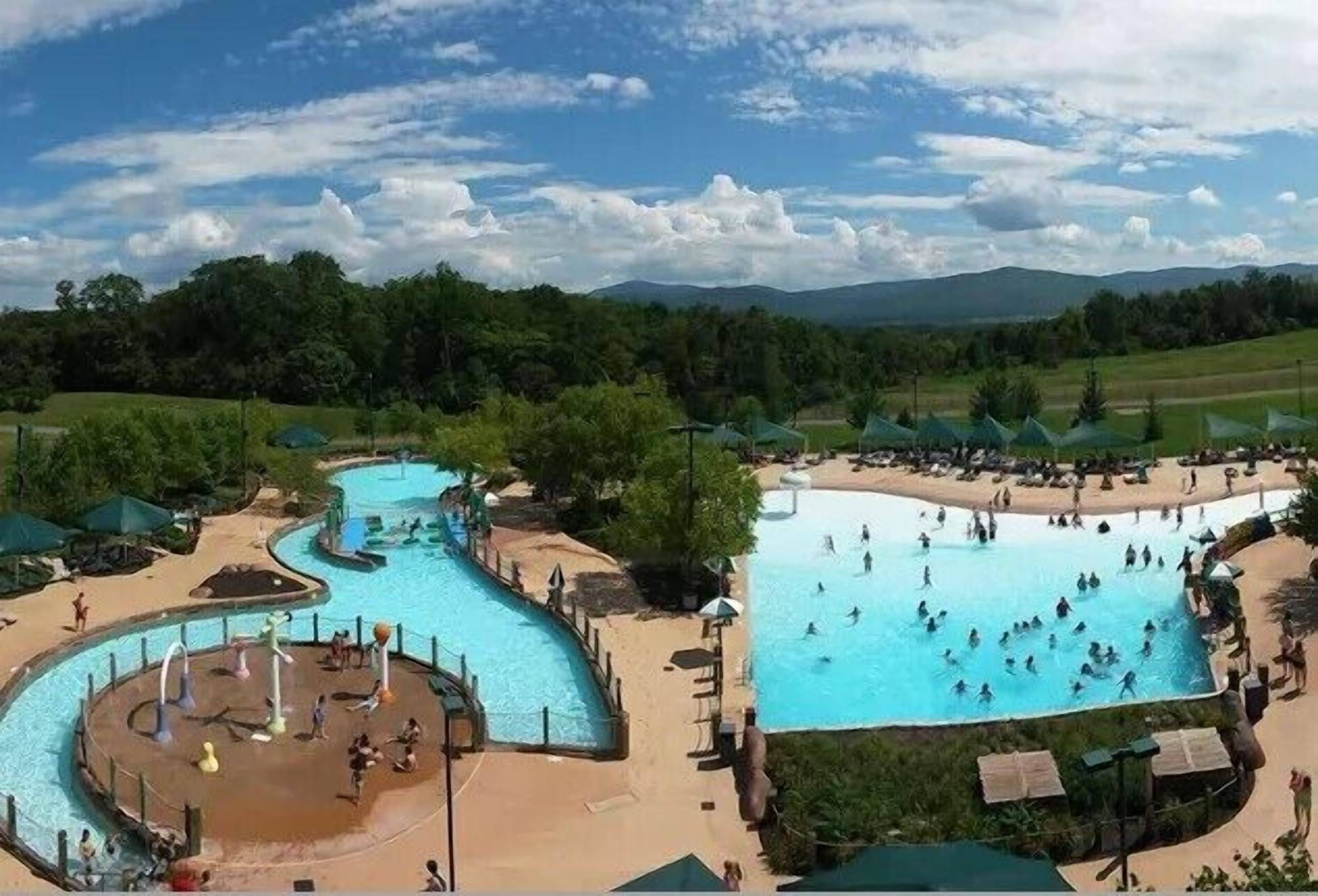 A large outdoor swimming pool with multiple lanes and a water slide, surrounded by umbrellas and lounge chairs.