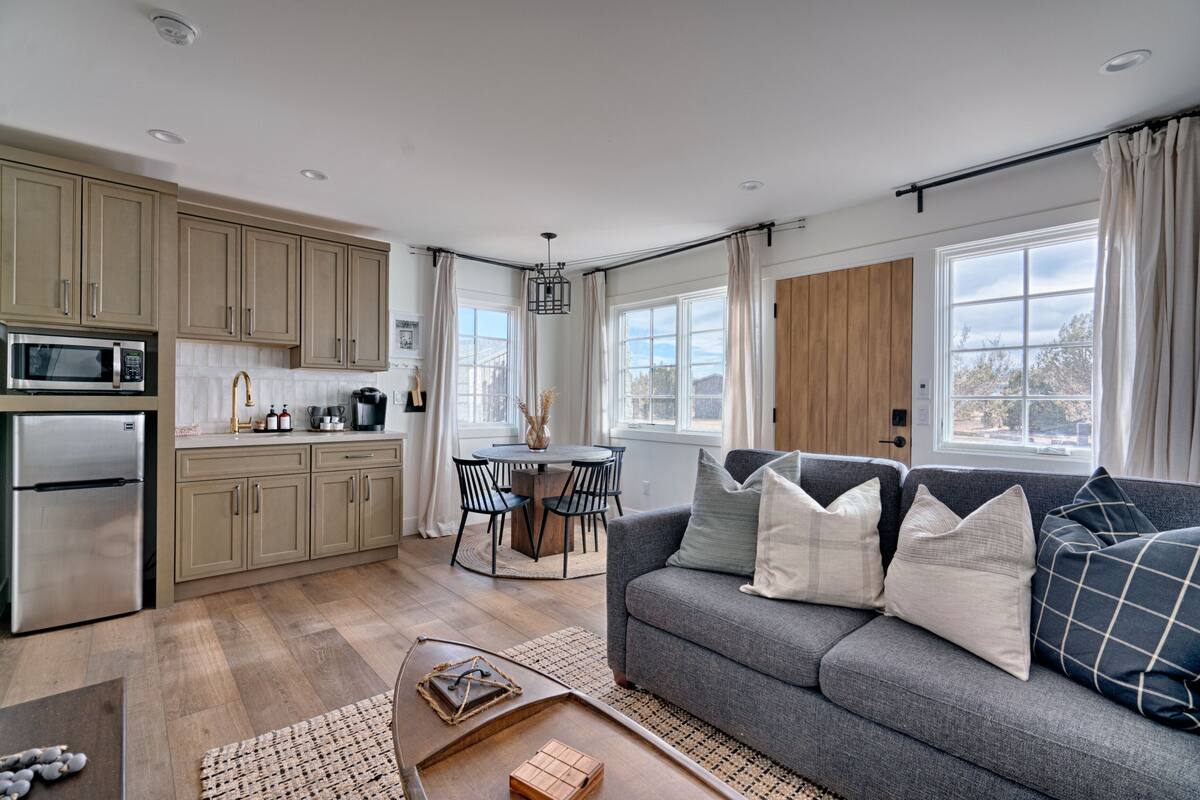 A modern kitchen with a dining area, a gray sofa with decorative pillows, and a wooden door.