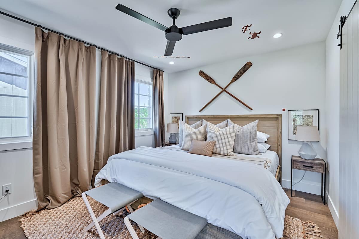 A bedroom with a bed, a ceiling fan, a window with curtains, and a wall-mounted lamp.