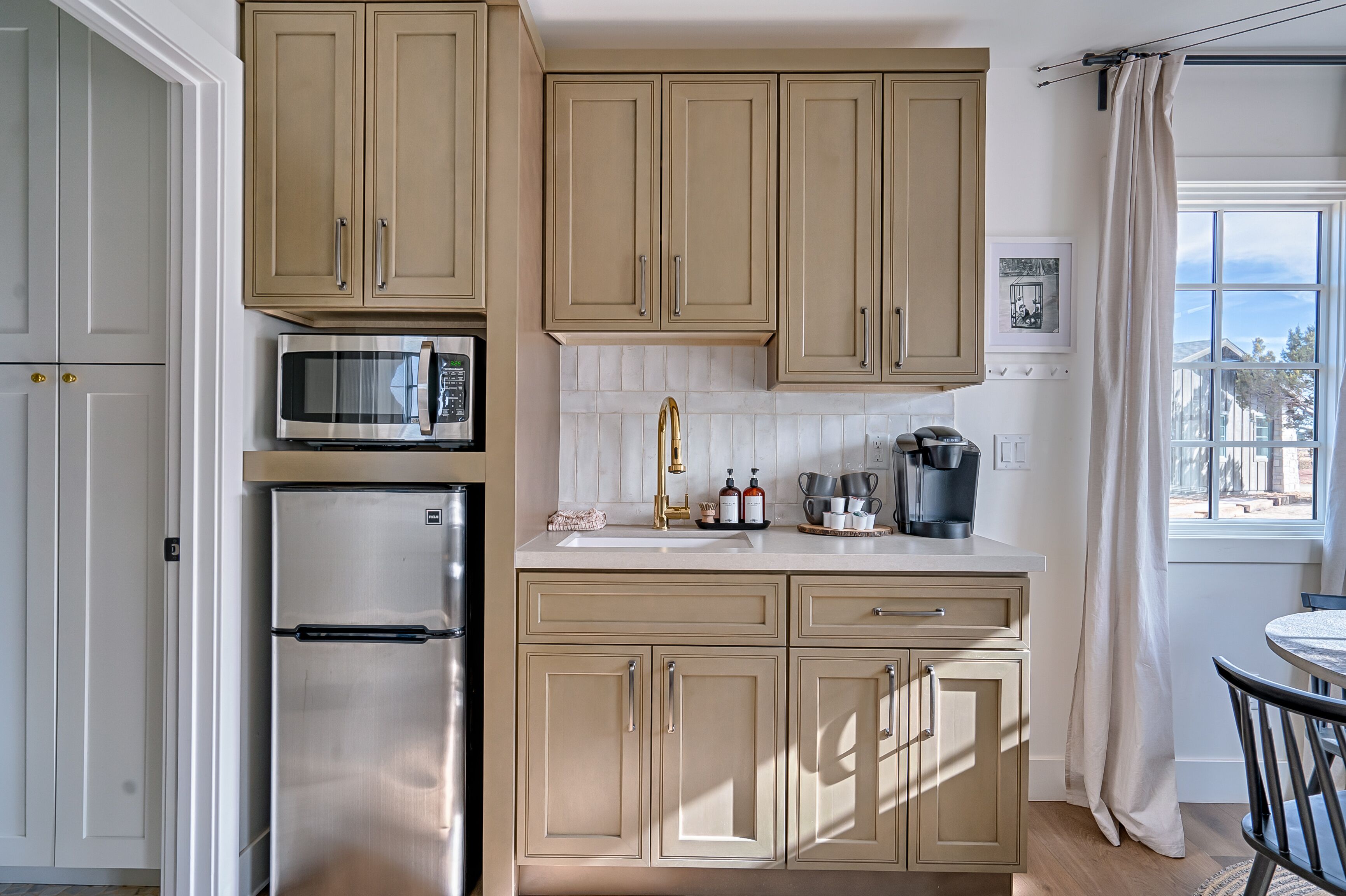 Een keuken met beige kastjes, een roestvrijstalen koelkast, een magnetron, een spoelbak en een raam met uitzicht op huizen.