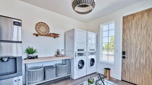 A modern laundry room with a stainless steel refrigerator, a washing machine, and a dryer. There is a wooden shelf with decorative items and a potted plant.