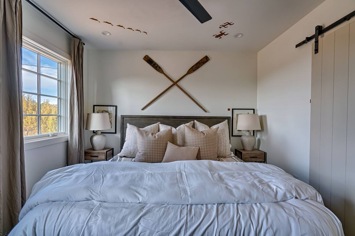 A bedroom with a large bed, two bedside tables with lamps, a headboard with a cross design, and a window with curtains.