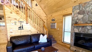A wooden interior with a staircase, a black leather sofa, a coffee table, and a stone fireplace. - Cozy cabin w/ loft, private hot tub, near Bison Ranch, sleeps 6 (Overgaard)