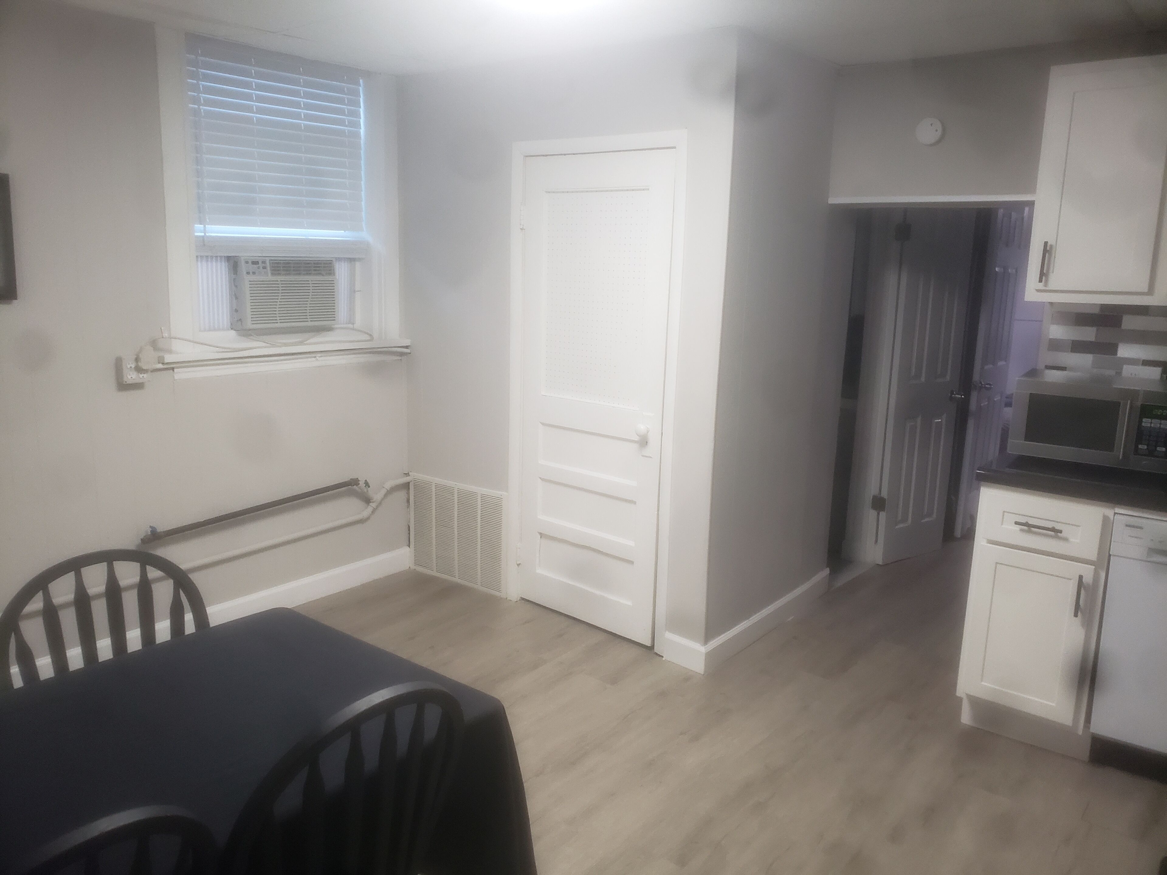 Una cocina pequeña y limpia con gabinetes blancos, un microondas y un lavavajillas. Hay un comedor con una mesa y sillas, y una puerta que da a otra habitación.