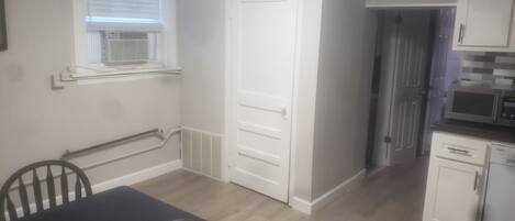 Una cocina pequeña y limpia con gabinetes blancos, un microondas y un lavavajillas. Hay un comedor con una mesa y sillas, y una puerta que da a otra habitación.