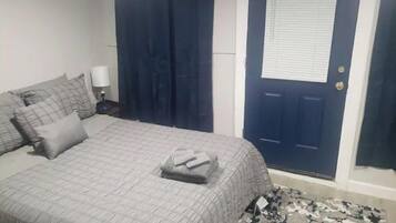 A bedroom with a bed, pillows, a nightstand, a lamp, a dark blue door, and a patterned rug.