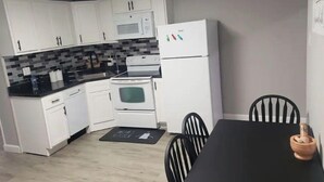 A compact kitchen with white cabinets, a black countertop, a microwave, an oven, and a refrigerator. There is a dining table with chairs and a mat on the floor.